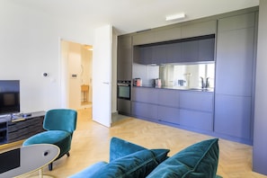 A modern kitchen with grey cabinets and a built-in oven. There is a teal armchair and a small table with a black surface. - Apartment Elizabeth - Les Demoiselles at Versailles (Versailles)