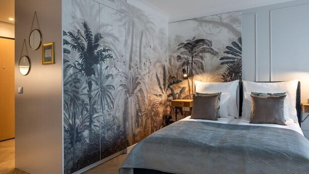 A bedroom with a large mural of tropical foliage on the wall, a bed with a grey patterned bedspread, and a bedside table with lamps.