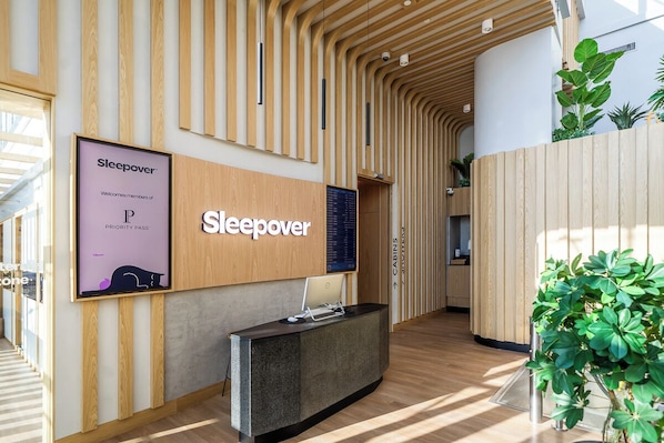 A reception area with a wooden reception desk, a large sign for Sleepover, and a digital display. - Sleepover Terminal 3, Concourse B – formerly sleep ‘n fly (Dubai)