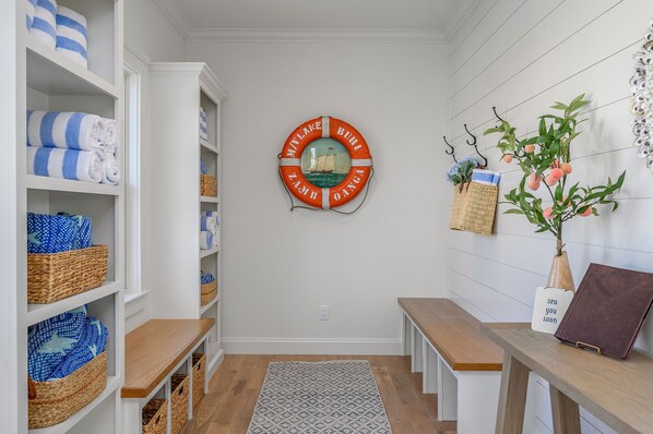 A hallway with white shelves holding towels and baskets, a life preserver decoration, a bench, and a small plant. - Bright & Beautiful Waterfront Cape Cod Home (Sandwich)