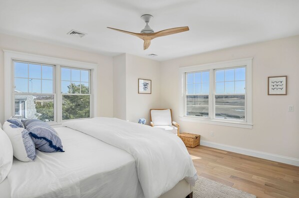 A bedroom with a large bed, a ceiling fan, two windows, and a chair. - Bright & Beautiful Waterfront Cape Cod Home (Sandwich)