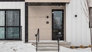 A building with a beige facade, black-framed windows, and a door with the number 705. Snow is falling, and there are steps leading to the entrance. - Wake Up to Bay Views | Walk Downtown + Beach (Traverse City)