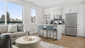 A modern kitchen with white cabinets, a stainless steel refrigerator, and a bar area with two chairs. - Wake Up to Bay Views | Walk Downtown + Beach (Traverse City)