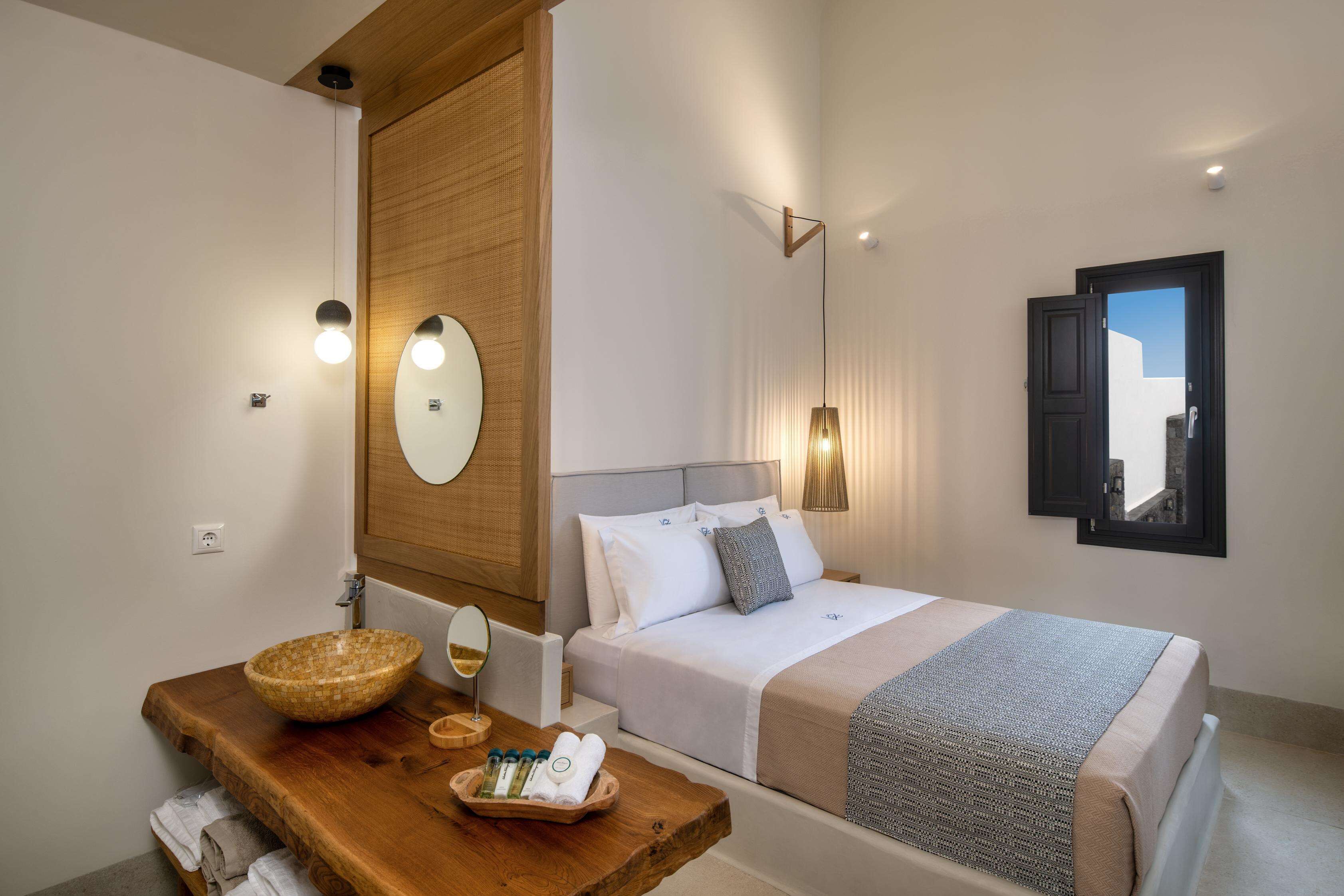 A bedroom with a wooden vanity, a large mirror, a bed with white linens, and a view of a building through a window.
