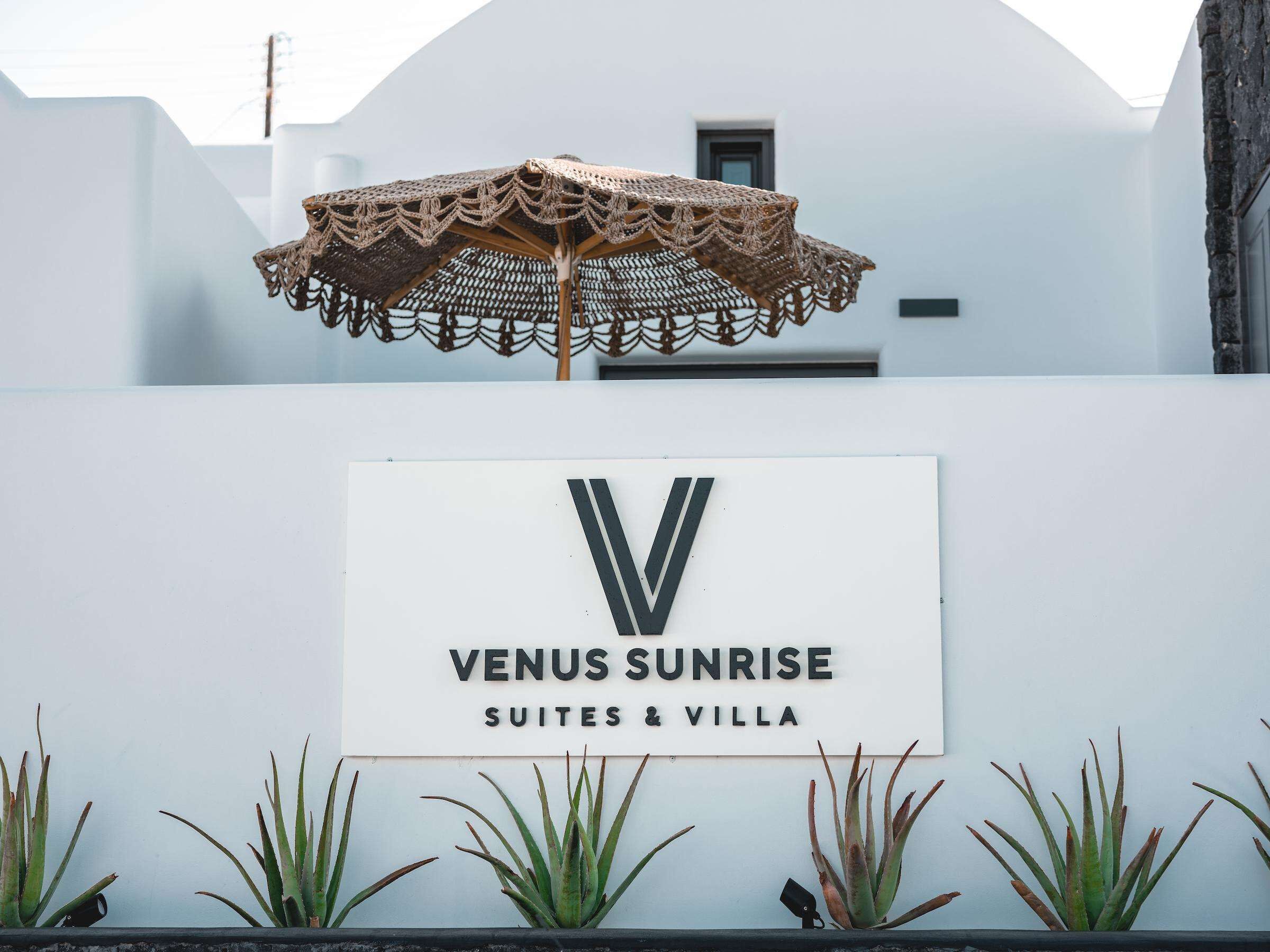 Une enseigne pour «Venus Sunrise Suites & Villa », avec un grand logo en forme de «V », entouré de plantes en pot.