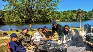 Un gruppo di persone riunite attorno a un tavolo all'aperto, con uno specchio d'acqua e alberi sullo sfondo.