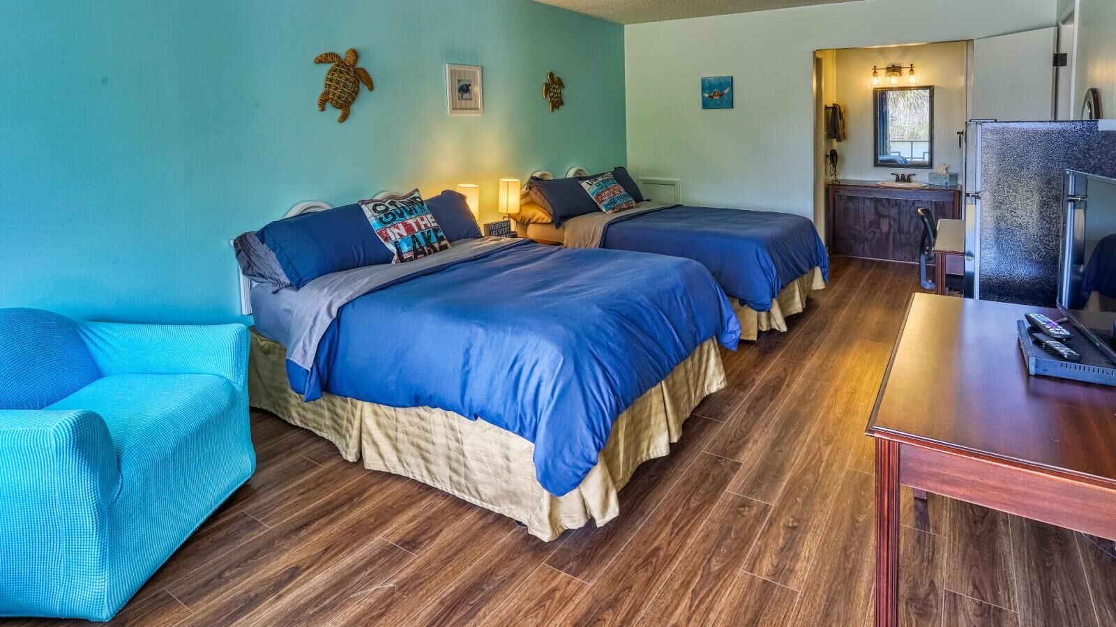 Habitación con dos camas, piso de madera, un pequeño sofá, un escritorio y un baño visible a través de una puerta abierta.
