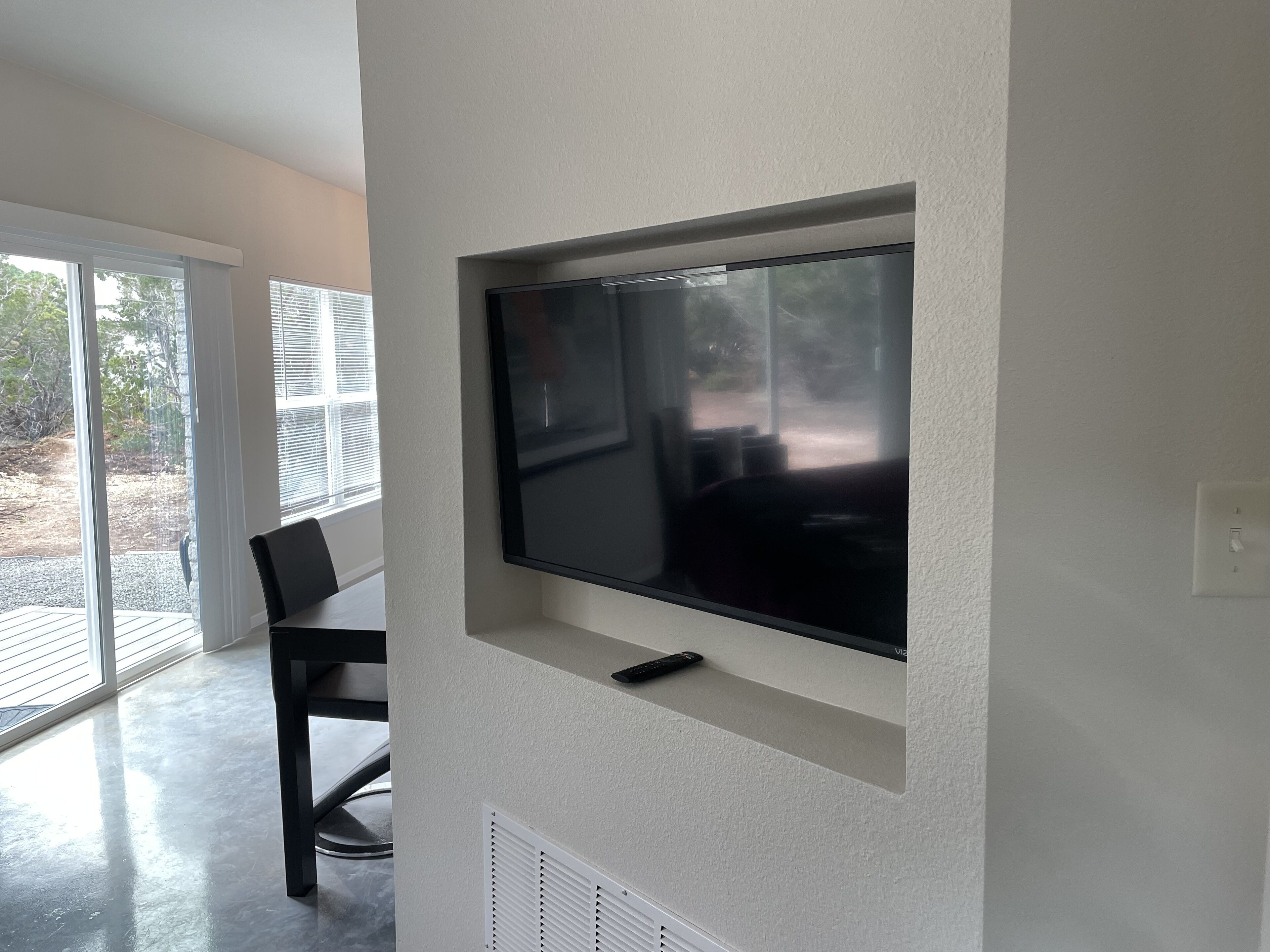 A modern living room with a flat-screen TV mounted on a white wall, a dining area with a table and chairs, and a sliding glass door leading to an outdoor patio.