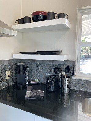 Uma cozinha moderna com bancada preta, uma cafeteira Keurig, uma torradeira e um conjunto de canecas pretas sobre uma estante branca.