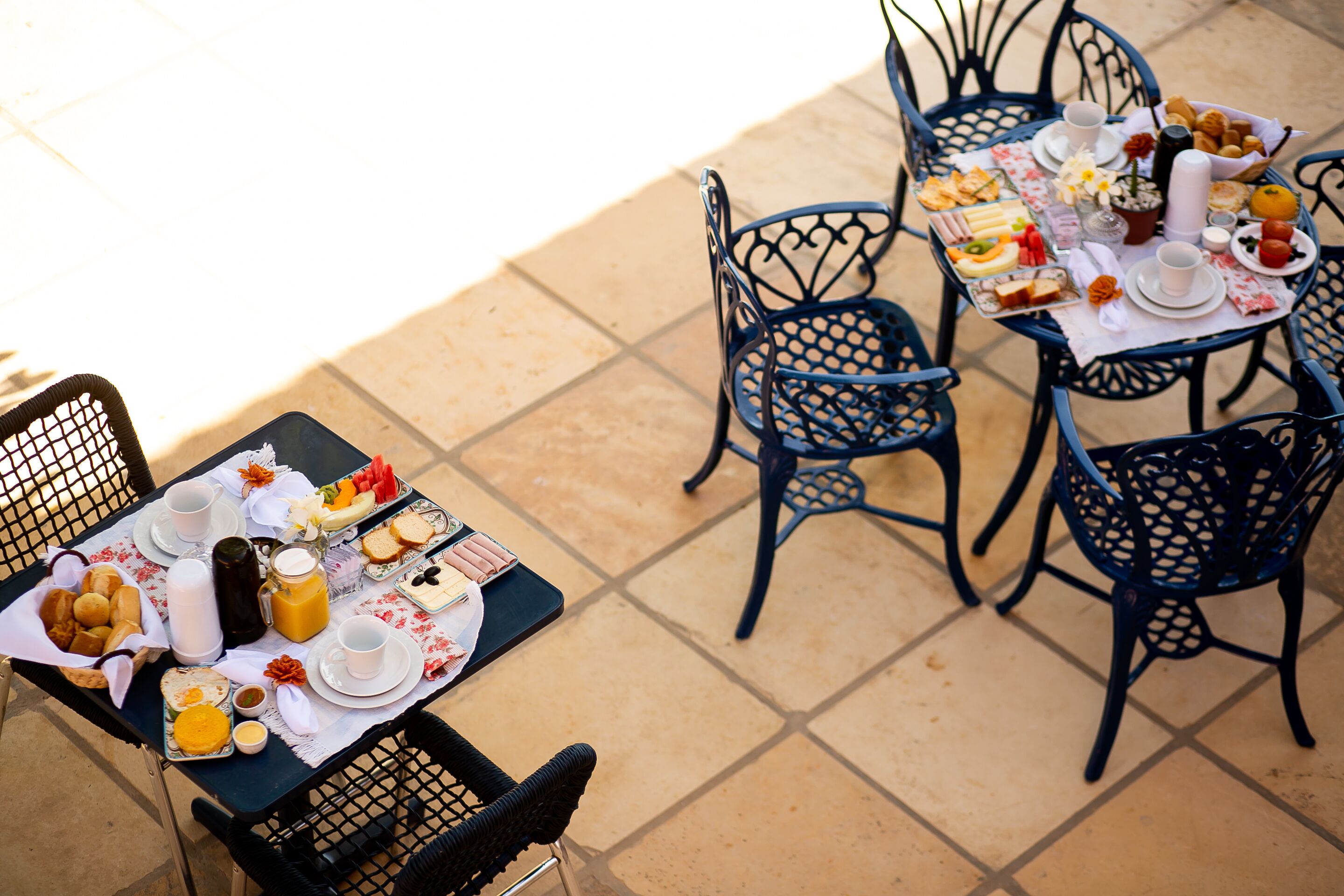 Une table dressée avec des aliments pour le petit-déjeuner, notamment du pain, des fruits et des boissons, entourée de chaises en métal sur une terrasse carrelée.