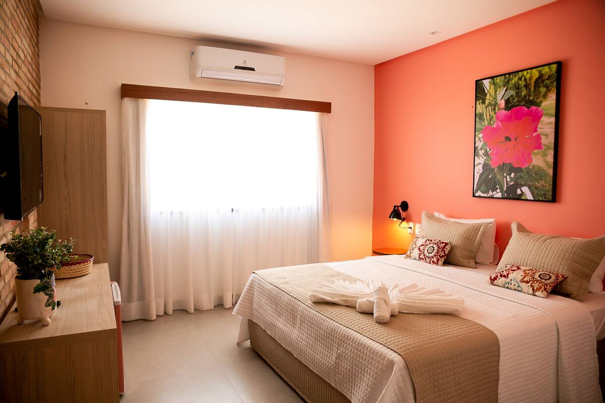 A hotel room with a bed, a window with curtains, a wall-mounted air conditioner, a bedside lamp, and a painting of a flower.