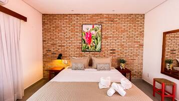 A hotel room with a brick wall, a large bed with multiple pillows, a bedside table with a lamp, and a red stool.