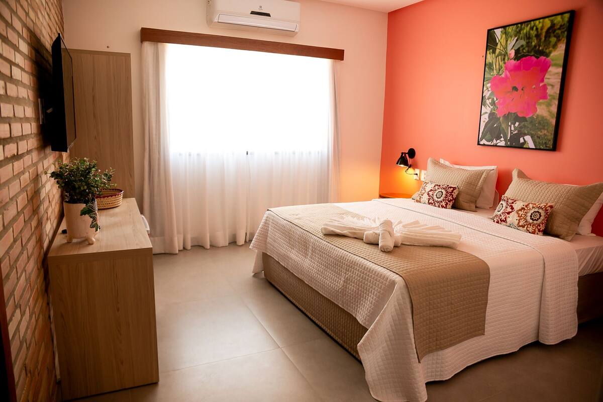 A hotel room with a bed, a bedside table, a potted plant, a window with curtains, and a wall-mounted air conditioner.