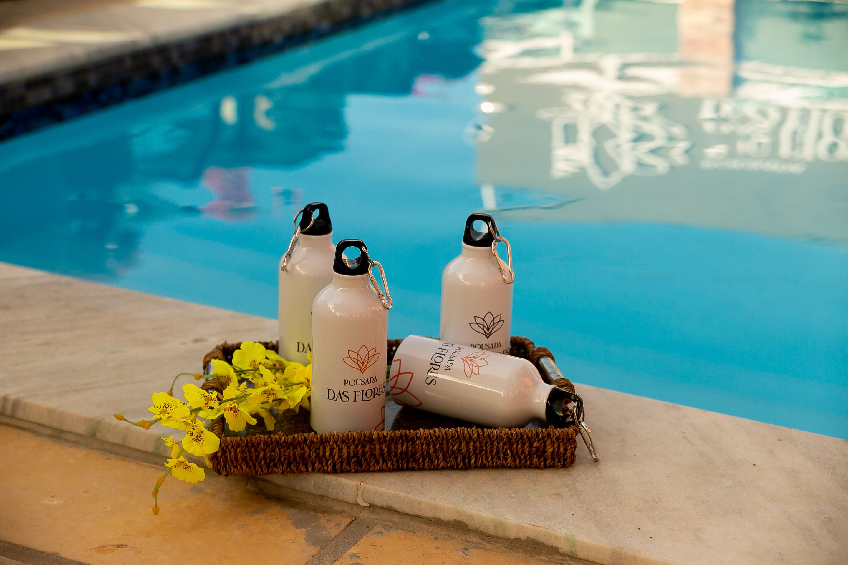 A poolside setting with three bottles labeled "Pousada DAS FLORES" and a basket of yellow flowers.