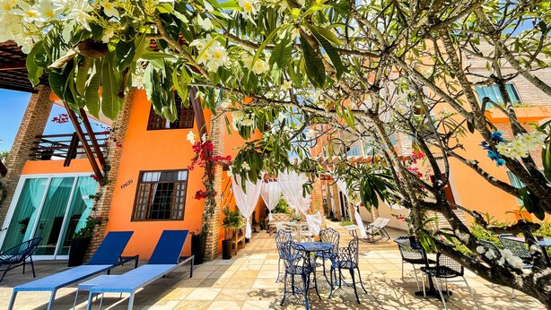 An outdoor seating area with lounge chairs and tables, surrounded by flowering trees and a building with orange walls. - Pousada das Flores (Luis Correia)