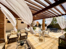 Un espacio al aire libre cubierto con mobiliario de mimbre y una mesa de vidrio, rodeado de cortinas blancas.