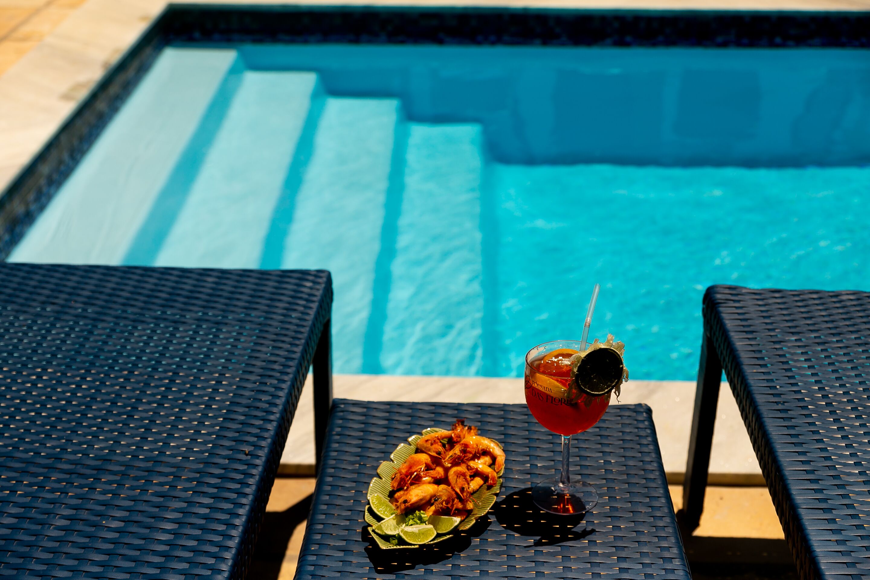 A poolside setting with a table holding a plate of food and a drink, and two lounge chairs.