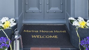 Une porte à deux battants avec un trou de serrure et une sonnette, un paillasson sur lequel est inscrit « Bienvenue à l’hôtel Marina House », et deux jardinières avec des fleurs blanches.