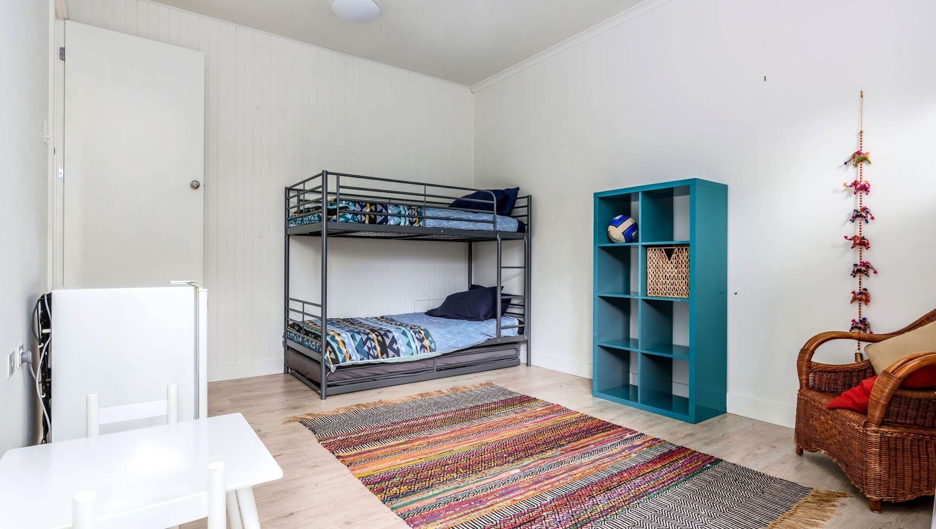 A bunk bed with patterned bedding, a teal shelving unit, a wicker chair, and a white table.