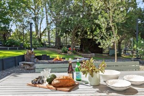 Un área de comedor al aire libre con una mesa de madera dispuesta para una comida, que incluye pan, una botella y un jarrón con ramas.