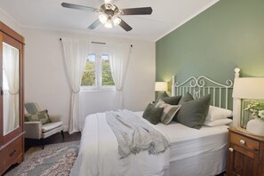 Une chambre à coucher comprenant un lit, une table de chevet, une chaise, un ventilateur de plafond et une fenêtre avec des rideaux.