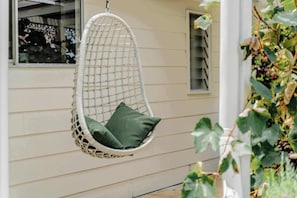 A white hanging chair with a green cushion in front of a house with beige siding. - Luxe Farm Stay: Animals, Fire Pit, Beach (Wamberal)