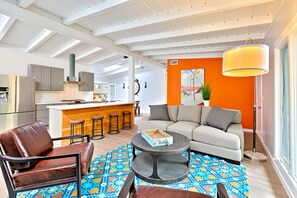 Una sala moderna con sofá, mesa de centro y sillas, una zona de cocina con taburetes, y una pared de acento anaranjada brillante.