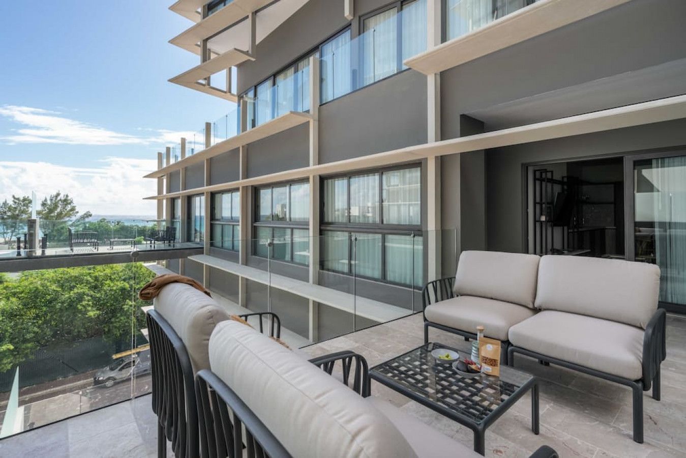 A balcony with a sofa and a glass table, overlooking a street and greenery.