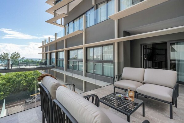 A balcony with a sofa and a glass table, overlooking a street and greenery.