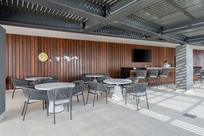 Um espaço interior moderno com várias mesas redondas e cadeiras, uma mesa de recepção e uma TV de tela plana na parede.