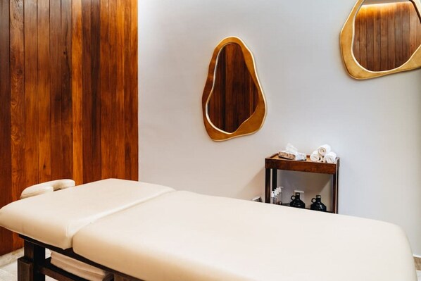 A treatment room with a wooden wall, a wooden-framed mirror, a small wooden table with toiletries, and a white examination table.