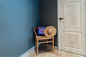 A wooden chair with a woven bag and a straw hat against a blue wall, next to a wooden door.