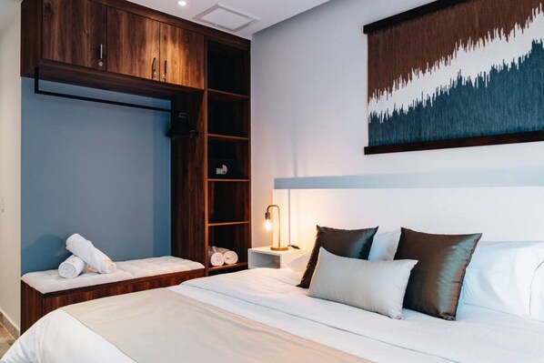 A hotel room with a bed, a bedside table with a lamp, a wardrobe, and a painting on the wall.