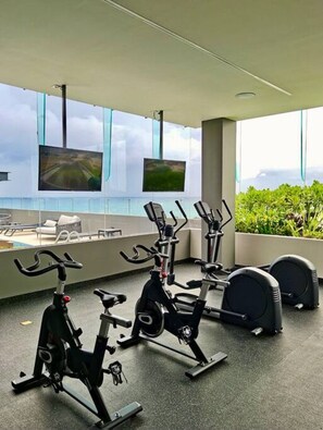 Un gimnasio con bicicletas estáticas y máquinas elípticas, amplias ventanas con vistas a zonas verdes e iluminación empotrada en el techo.