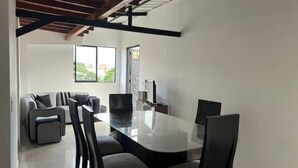 Un comedor moderno con una mesa y sillas de vidrio, un conjunto de sofás y un ventanal con vistas al verde exterior.