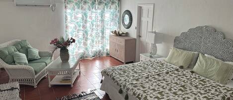 Un dormitorio con cama, una silla, una mesita con un florero, un tocador y una ventana con cortinas.