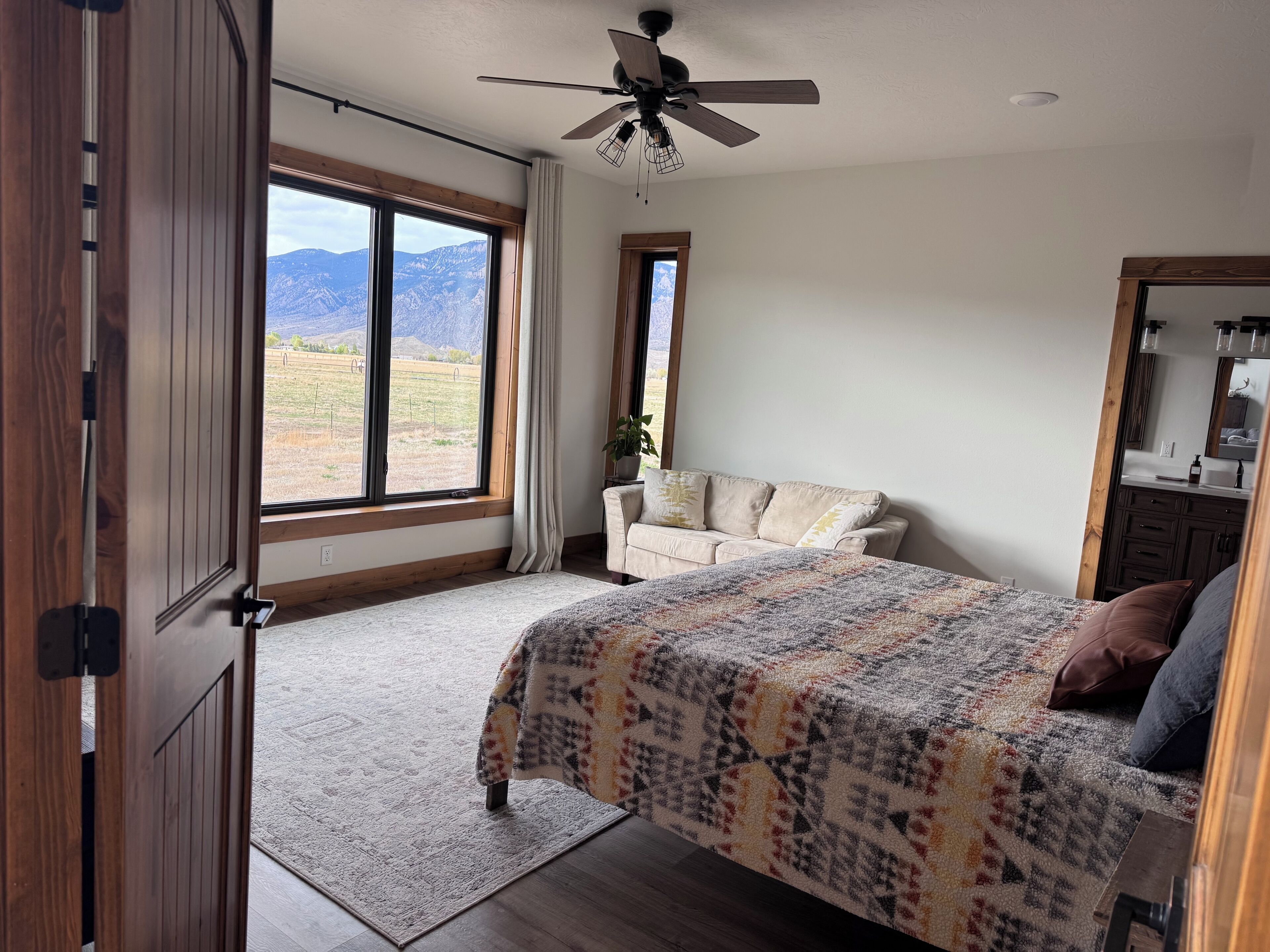 A bedroom with a large window, a bed, a ceiling fan, and a wooden wardrobe.