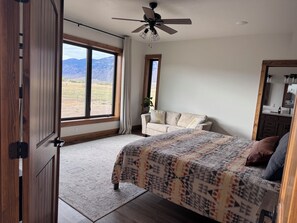 A bedroom with a large window, a bed, a ceiling fan, and a wooden wardrobe. - The Rendezvous - Mountain views surrounded by fields (Cody)