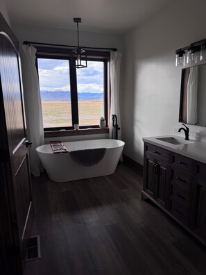 A bathroom with a freestanding bathtub, a window with a view of mountains, and a wooden vanity. - The Rendezvous - Mountain views surrounded by fields (Cody)
