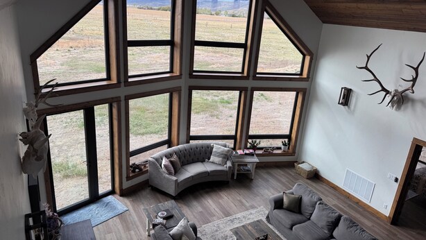 A spacious living room with large windows, a curved gray sofa, a wooden coffee table, and antler wall decor. - The Rendezvous - Mountain views surrounded by fields (Cody)