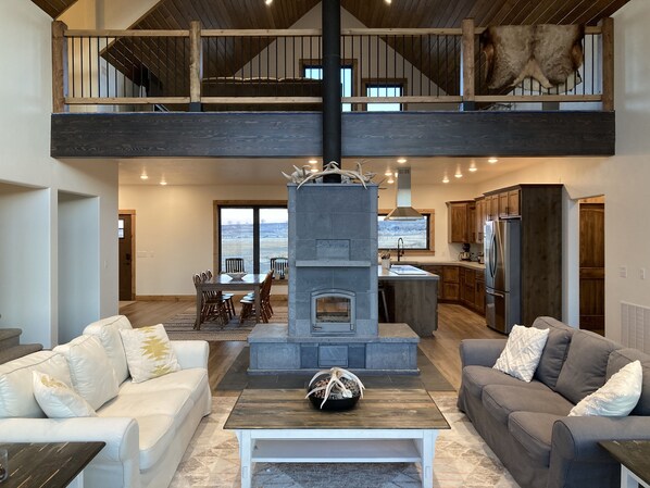 A spacious living area with a fireplace, two sofas, a dining table, and a kitchen in the background. - The Rendezvous - Mountain views surrounded by fields (Cody)
