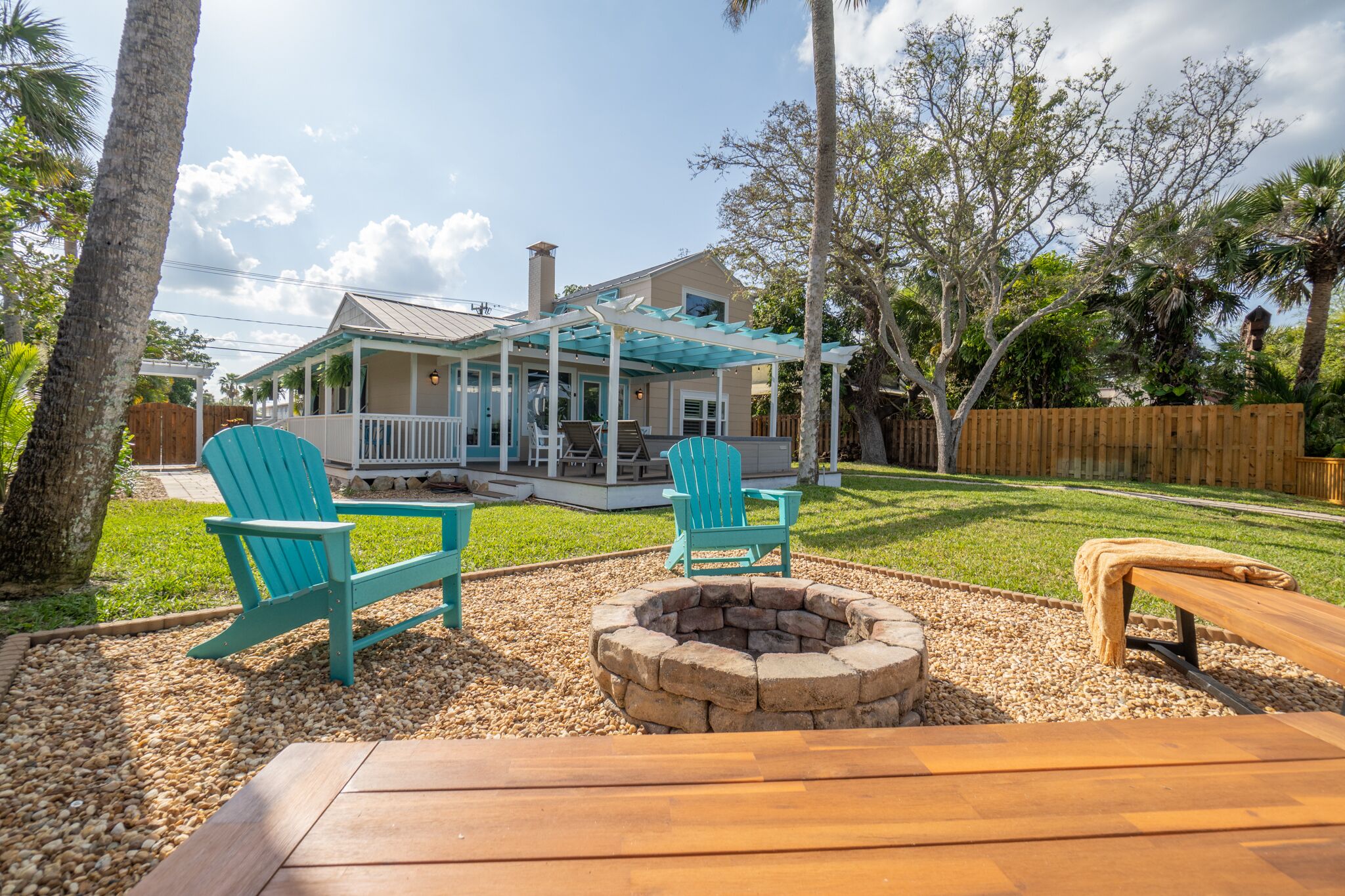 A house with a covered patio, two turquoise Adirondack chairs, a stone fire pit, and a wooden bench.