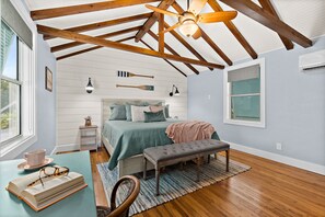 Une chambre avec un plafond en bois, un lit recouvert d’une couverture verte, un banc et une petite table sur laquelle se trouvent un livre et des lunettes.