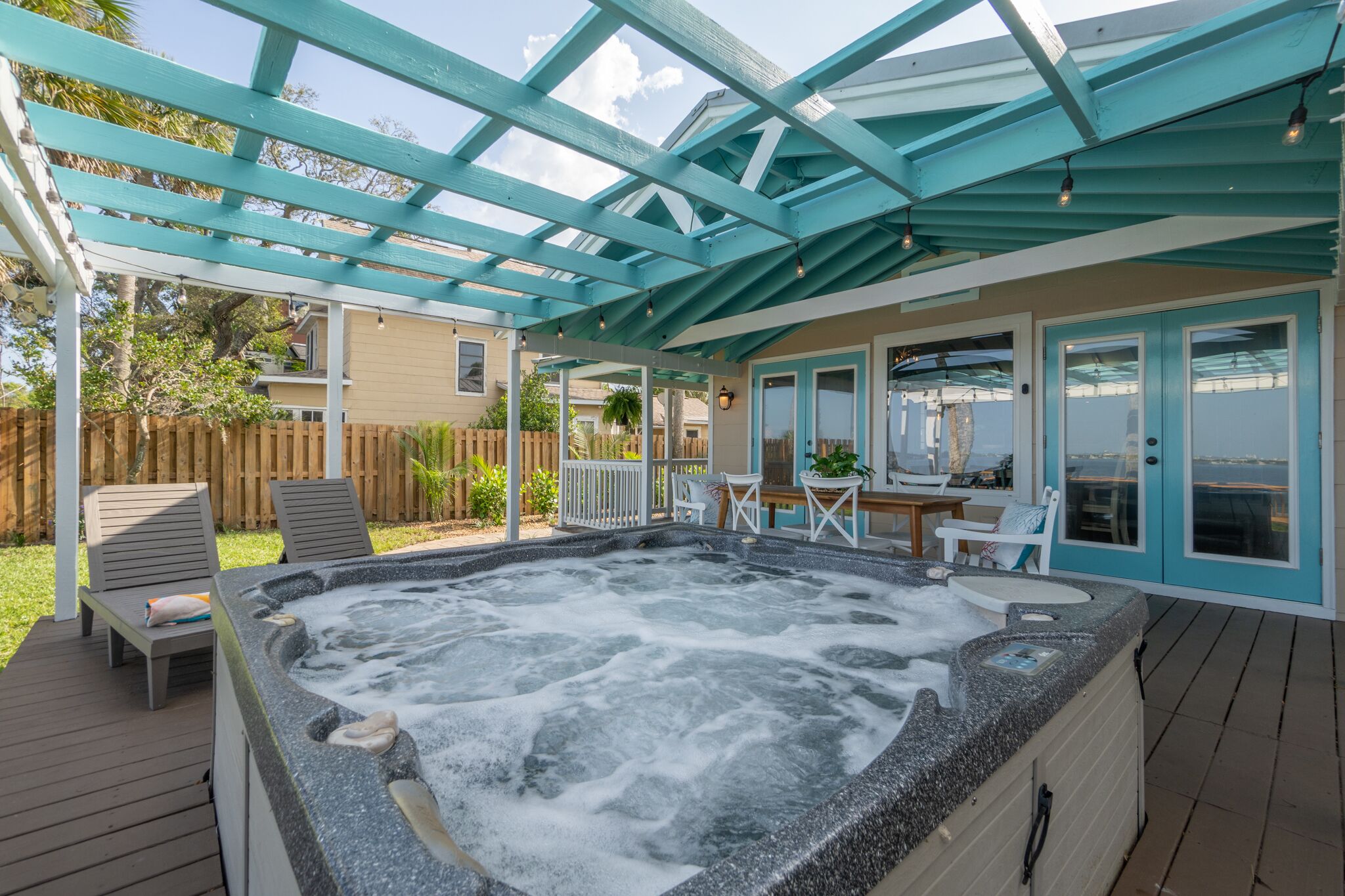 Ein Whirlpool unter einer blauen Pergola mit einer Holzterrasse, Sitzgelegenheiten im Freien und Blick auf eine Wohngegend.