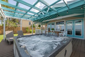 Un bain à remous sous une pergola bleue, avec une terrasse en bois, des sièges d’extérieur et une vue sur un quartier résidentiel.
