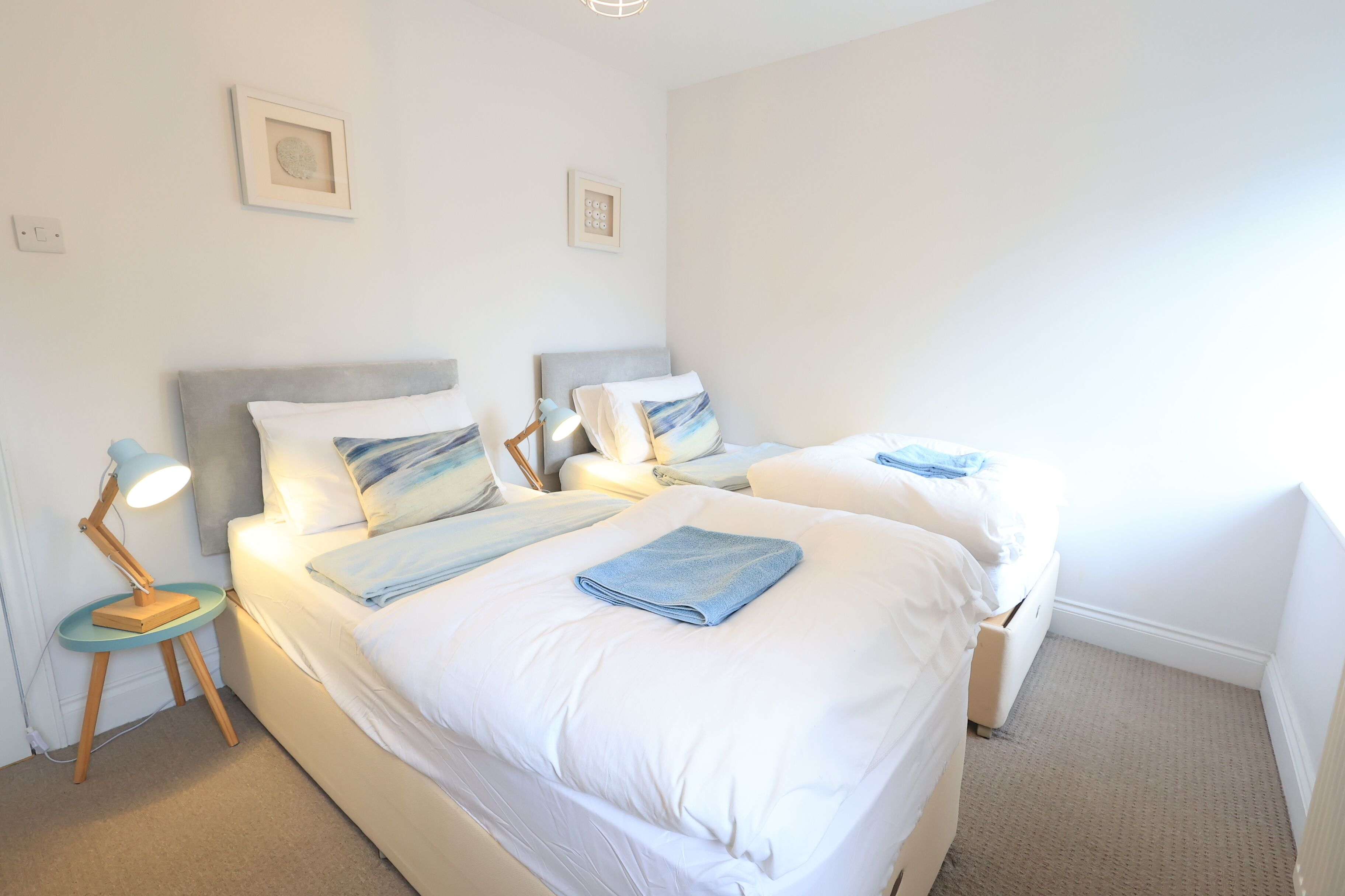 A bedroom with two beds, each with a pillow and a folded towel. There is a small table with a lamp and a picture frame on the wall.
