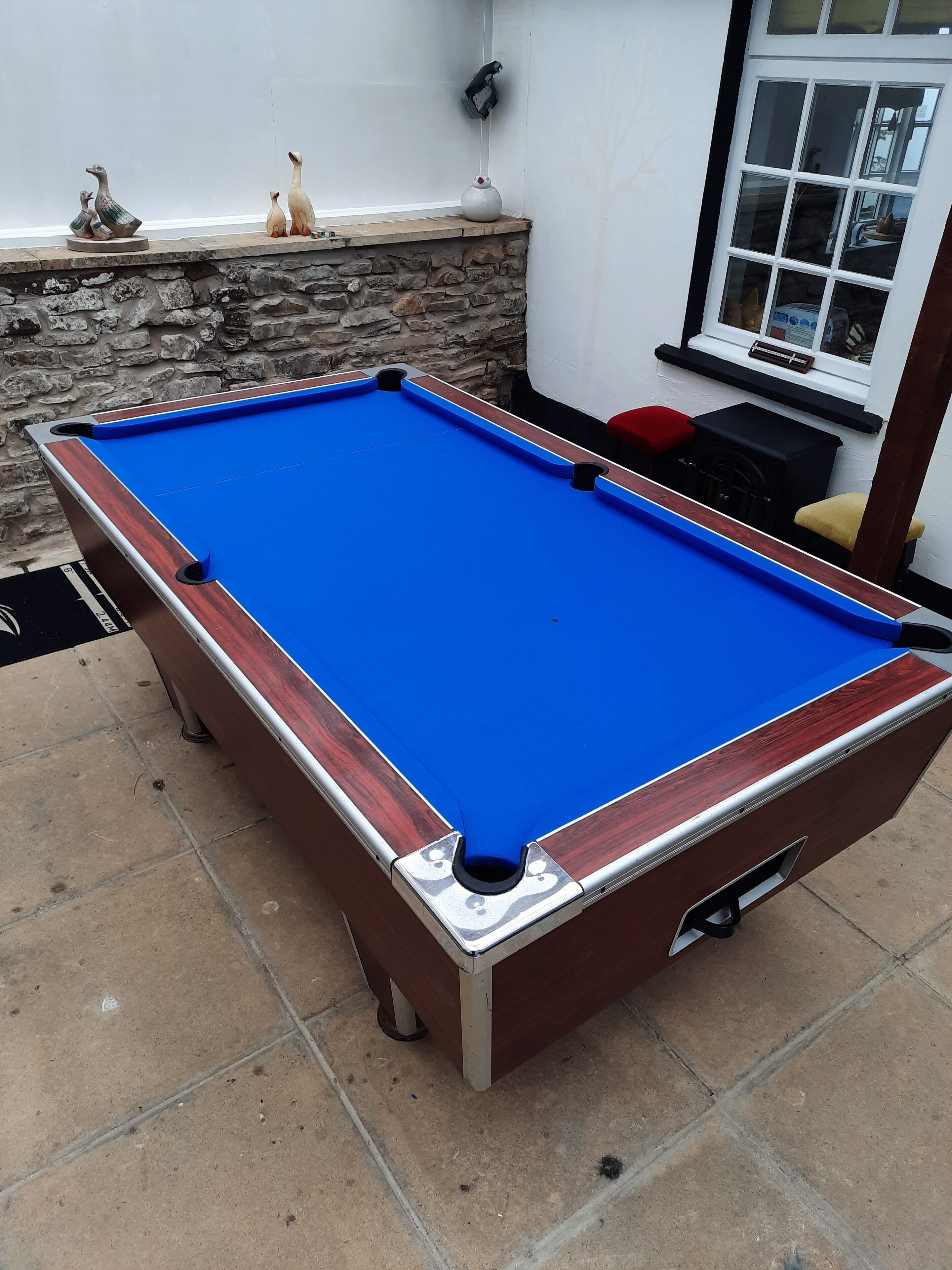 Una mesa de billar con superficie de fieltro azul, rodeada de paredes de piedra y una ventana.