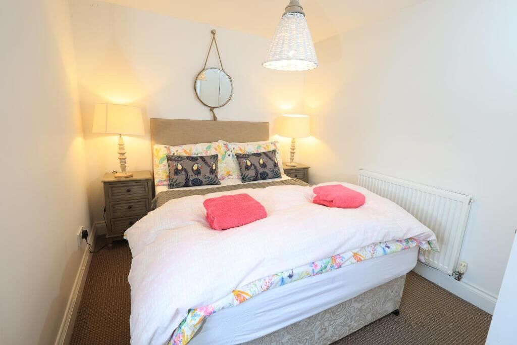A neatly made bed with white bedding and pink towels, a round mirror on the wall, and two bedside lamps.