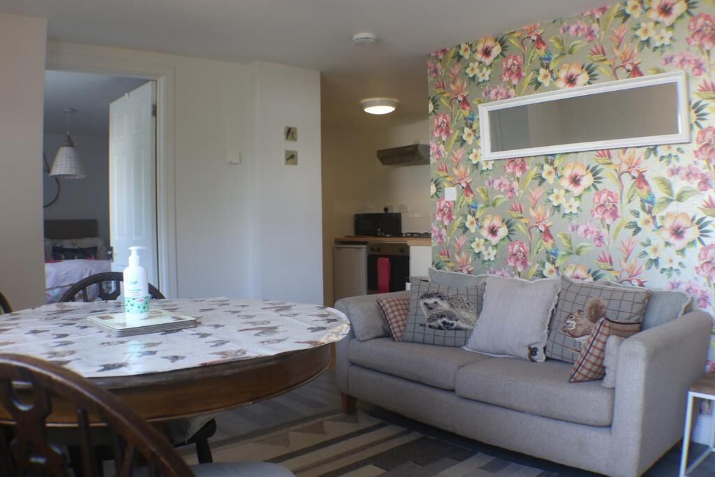A dining area with a round table, chairs, a sofa with decorative pillows, a wall-mounted mirror, and a floral wallpaper.