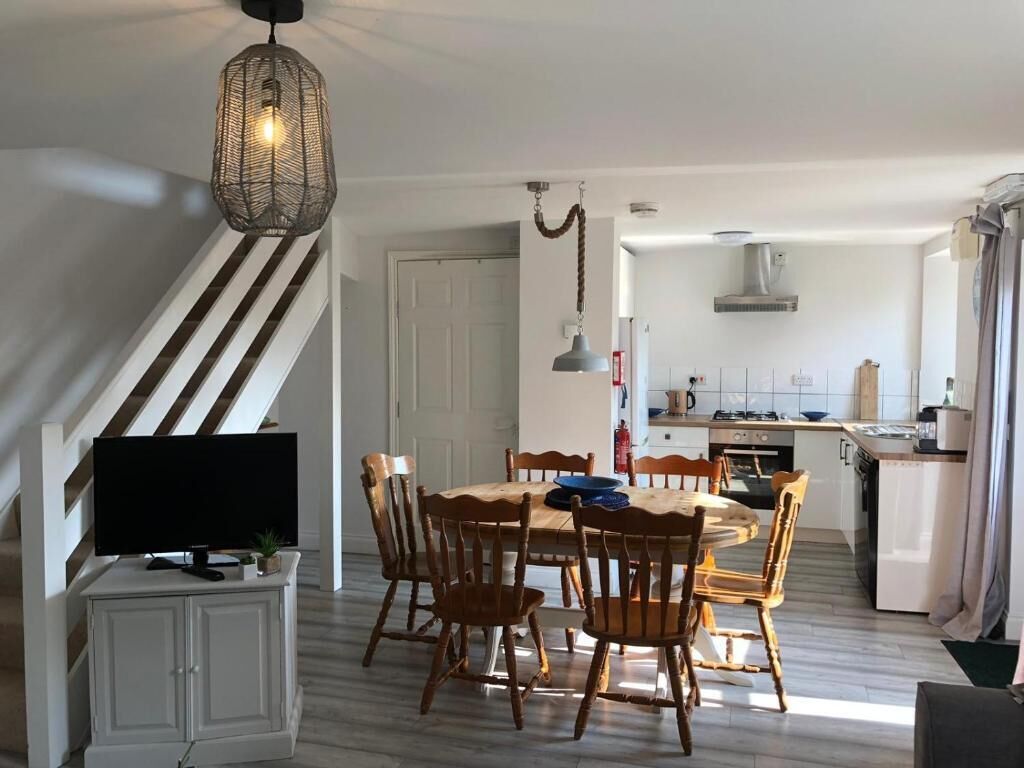 A dining area with a wooden table and chairs, a television on a cabinet, and a kitchen in the background.